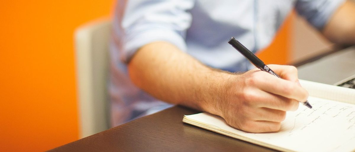 Kompetenzforum Unternehmensführung - eine kostenlose Info-Veranstaltung für Arbeitgeber im Handwerk. Eine Männerhand hält einen Stift über einem aufgeschlagenen Heft. Im Hintergrund ist ein aufgeschlagenes Notebook zu sehen.