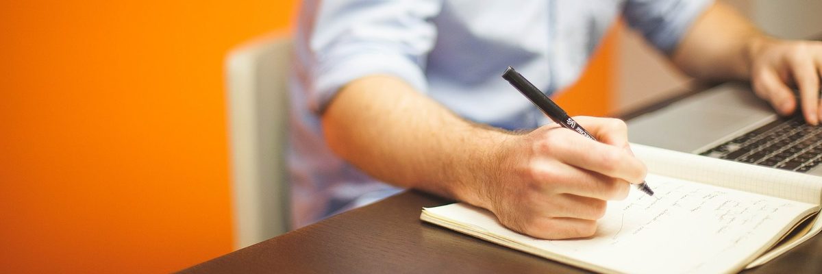 Kompetenzforum Unternehmensführung - eine kostenlose Info-Veranstaltung für Arbeitgeber im Handwerk. Eine Männerhand hält einen Stift über einem aufgeschlagenen Heft. Im Hintergrund ist ein aufgeschlagenes Notebook zu sehen.