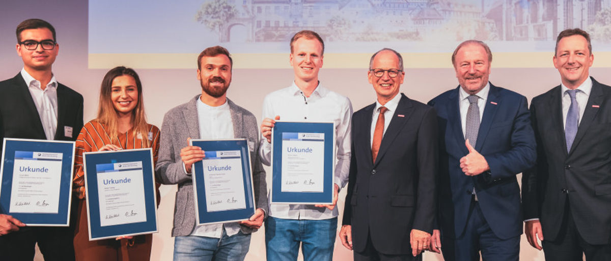 1. Sieger Simon Weber, Johannes Staudenmaier, Buket Yadak, Luca Härle. Vier junge Menschen halten gerahmte Urkunden vor sich, daneben stehen drei Herren in Anzügen. Alle lächeln in die Kamera.