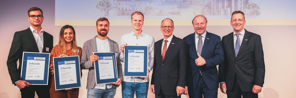 1. Sieger Simon Weber, Johannes Staudenmaier, Buket Yadak, Luca Härle. Vier junge Menschen halten gerahmte Urkunden vor sich, daneben stehen drei Herren in Anzügen. Alle lächeln in die Kamera.