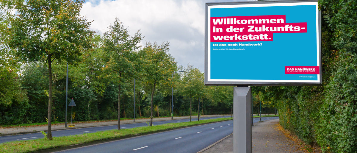 Die Imagekampagne macht deutschlandweit auf das Handwerk aufmerksam. Eine Werbetafel steh am Rand einer vierspurigen Straße mit Bäumen in der Mitte. Darauf ist ein blau-rot-weißes Werbeplakat.