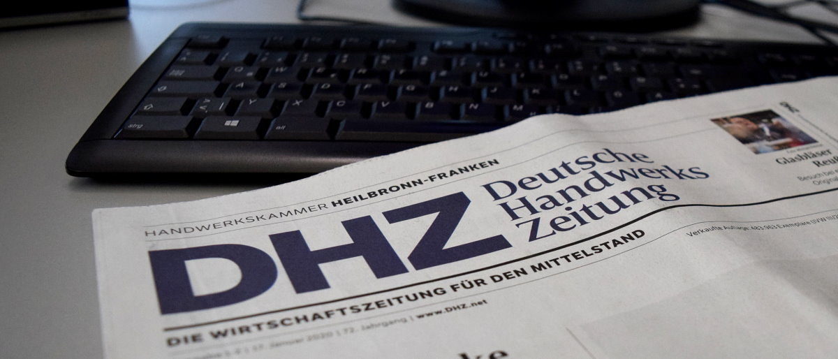 Die neue Ausgabe der Deutschen Handwerks Zeitung (DHZ) ist erschienen. Die obere linke Ecke einer Zeitung ist zu sehen. Sie liegt auf einem Schreibtisch. Im Hintergrund sieht man eine Tastatur.