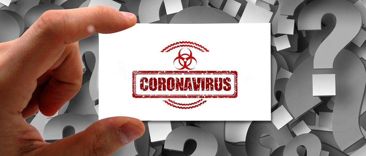 Eine Hand hält mit Daumen und Zeigefinger eine weiße Karte mit der Aufschrift Coronavirus. Im Hintergrund graue Fragezeichen,