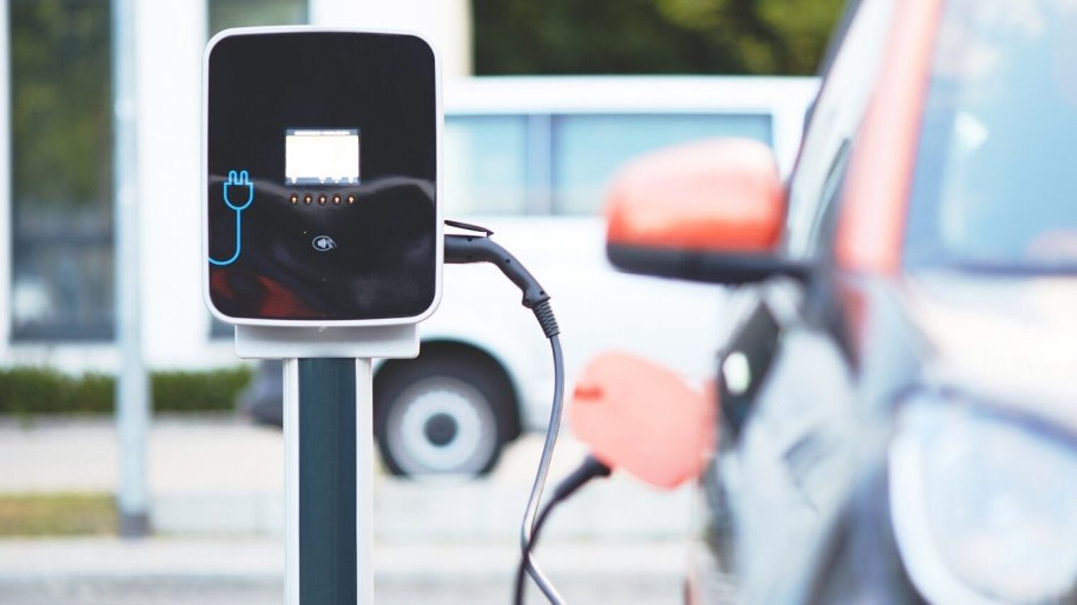 Eine moderne Ladesäule für Elektrofahrzeuge steht auf einem Parkplatz neben einem Auto, das gerade über das Kabel auflädt.