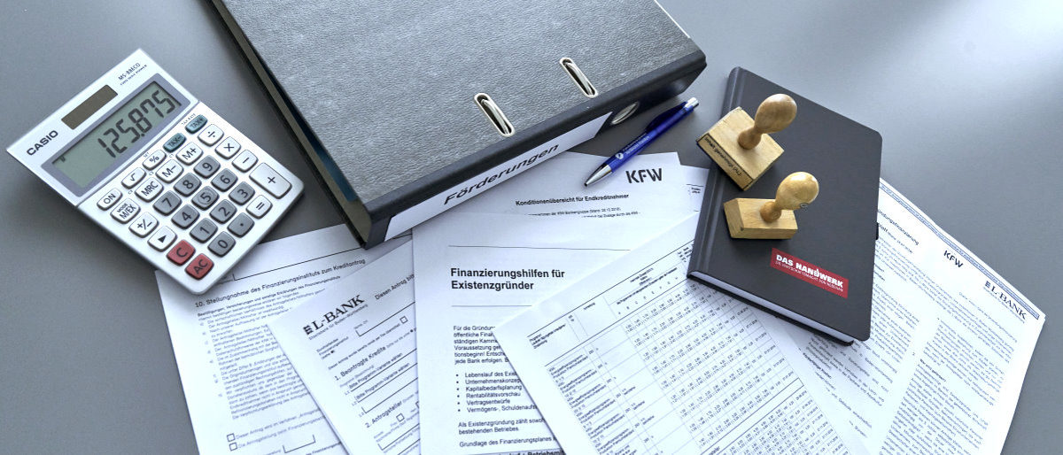Corona-Hilfsprogramme für betroffene Betriebe Papierformulare, ein Ordner, ein Taschenrechner, ein Notizbuch, zwei Stempel und ein Kugelschreiber liegen auf grauem Tisch.
