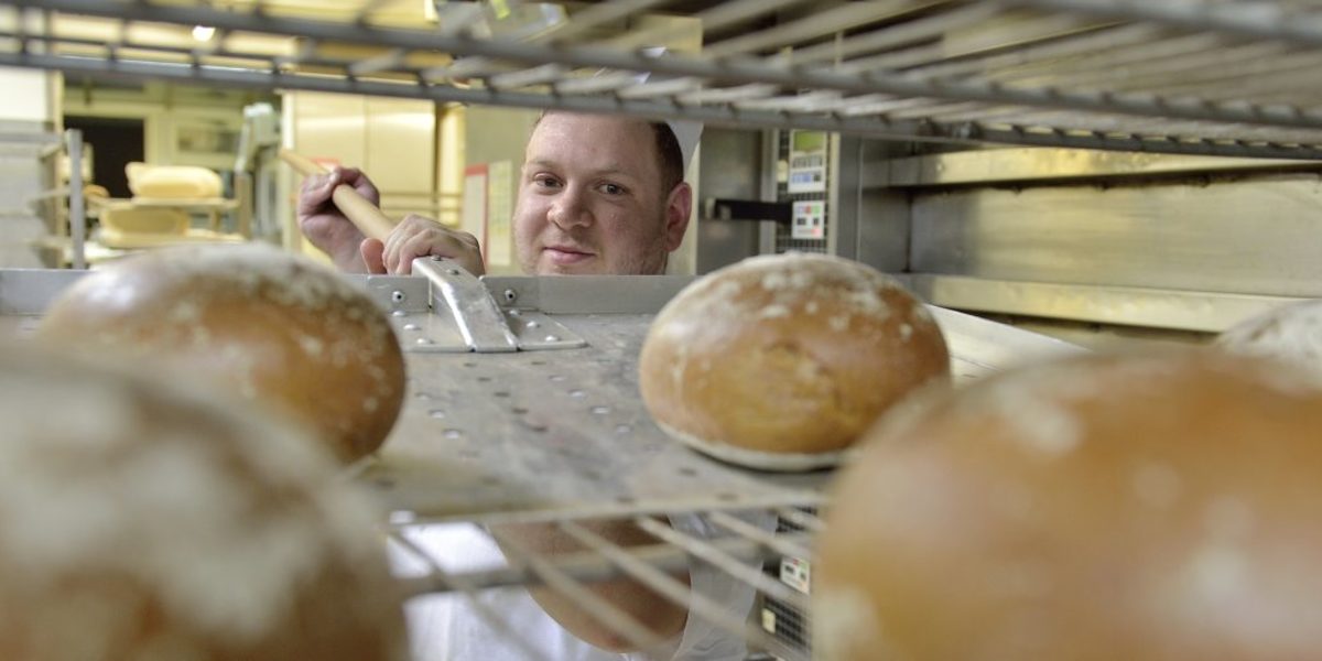 Brot vom Bäcker ist auch in Krisenzeiten noch gefragt. Ein Bäcker legt mit einem großen Schieber fertig gebackene Brote aus dem Ofen auf ein Gitterregal zum Abkühlen.