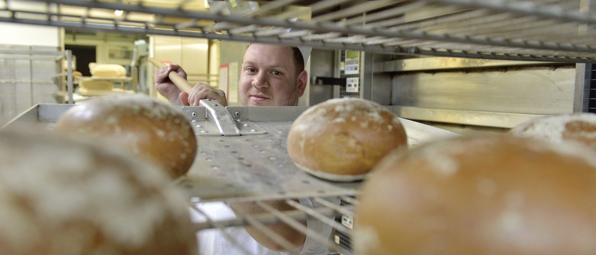Ein Bäcker legt mit einem großen Schieber fertig gebackene Brote aus dem Ofen auf ein Gitterregal zum Abkühlen.
