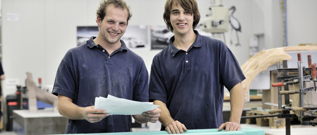 Zwei junge Männer stehen nebeneinander in einer Werkstatt und lächeln in die Kamera. Einer hält mehrere Blätter Papier.