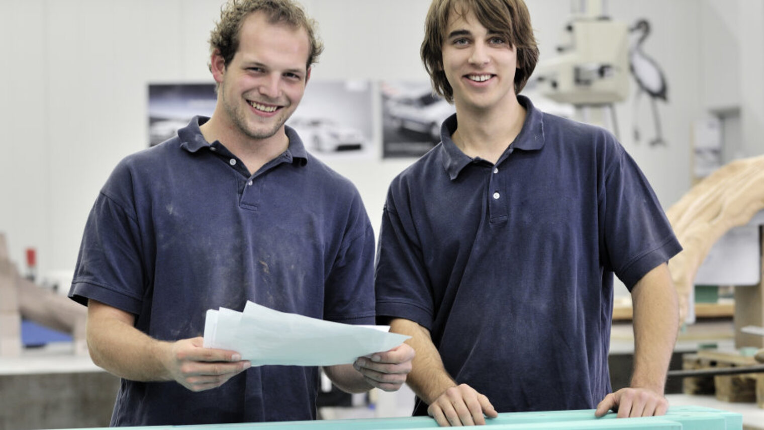 Zwei junge Männer stehen nebeneinander in einer Werkstatt und lächeln in die Kamera. Einer hält mehrere Blätter Papier.