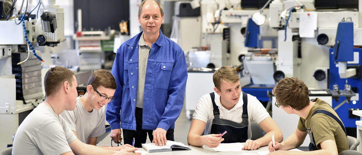 Betriebe erhalten für ihr Engagement in der Ausbildung eine Prämie . Vier junge Männer sitzen an einem Tisch und sprechen miteinander. Ein älterer Mann steht am Tisch und blickt in die Kamera.