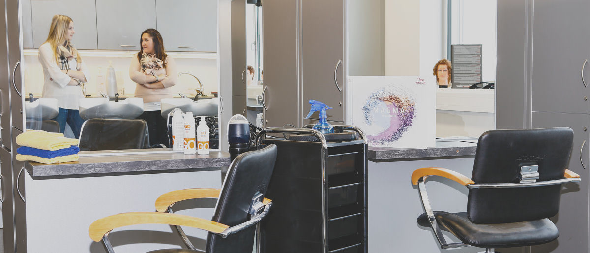 Die Corona-Krise hat vor allem Friseure wirtschaftlich hart getroffen. Zwei unbesetzte Stühle in einem Friseursalon. Im Spiegel sind zwei Frauen zu sehen, die Arme verschränkt, im Gespräch.