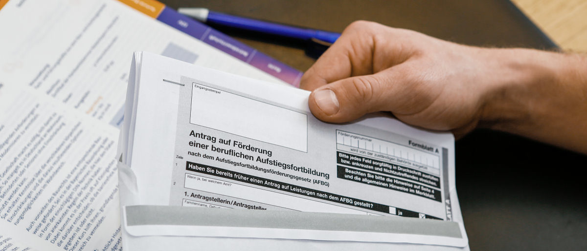 Für viele berufliche Weiterbildungen gibt es auch Förderprogramme. Eine Hand steckt auf einem Schreibtisch ein Antragsformular auf Ausbildungsförderung in einen Briefumschlag.