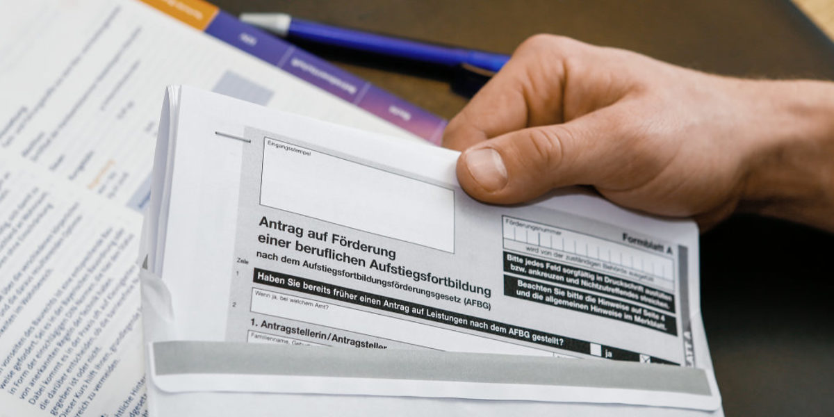 Eine Hand steckt auf einem Schreibtisch ein Antragsformular auf Ausbildungsförderung in einen Briefumschlag.