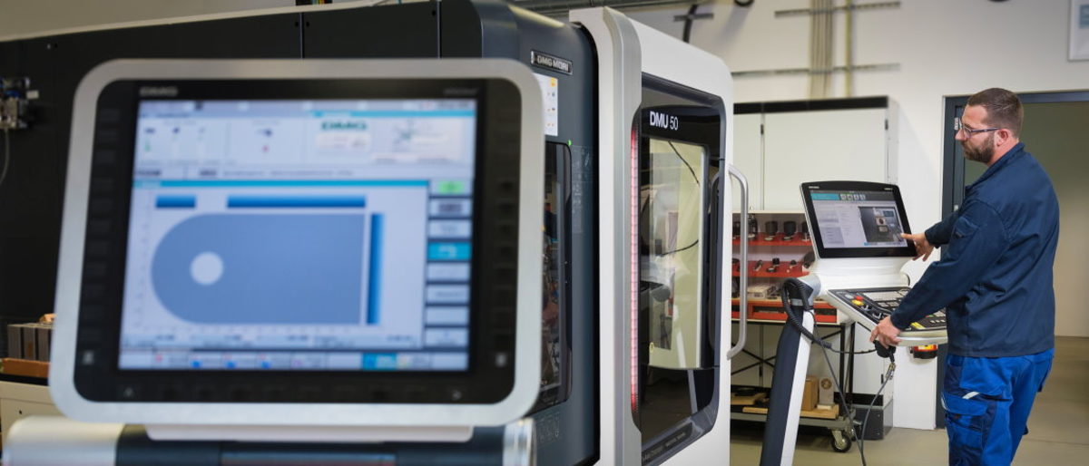 Digitalisierungsmaßnahmen werden gefördert. Im Vordergrund befindet sich ein Display einer CNC-Maschine. Im Hintergrund steht ein junger Mann an einem weiteren Display.