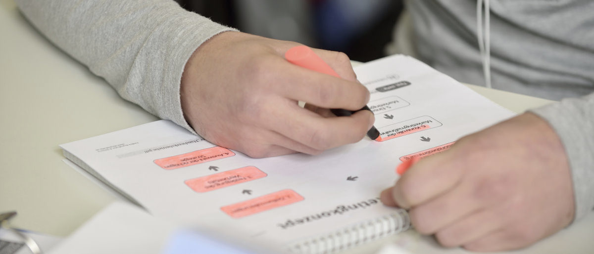 Meisterkurse bereiten Teilnehmer gezielt auf die Meisterprüfung vor. Zwei Hände, ein Block mit Arbeitsblatt auf einem Tisch. Die rechte Hand markiert einen Textabschnitt mit rotem Textmarker.