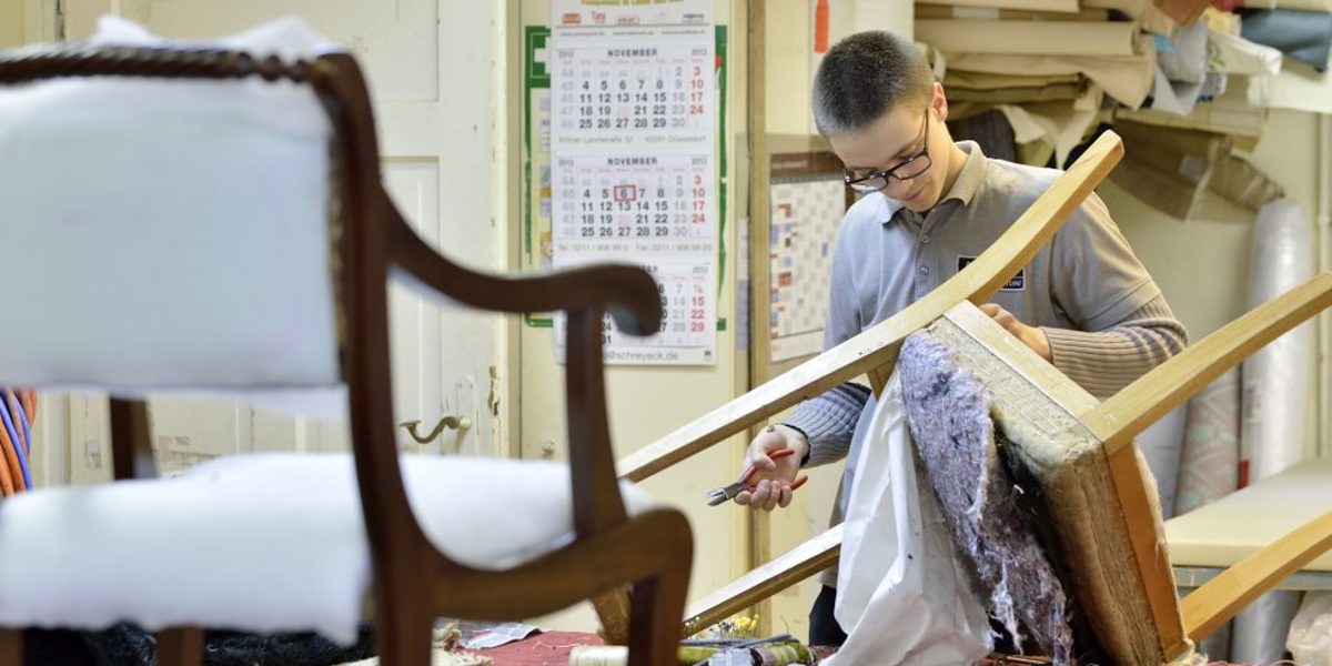 Raumausstatter gehören seit 2020 wieder zum zulassungspflichtigen Handwerk. Im Vordergrund steht erhöht ein Stuhl. Weiter hinten bezieht ein junger Mann einen Stuhl neu.
