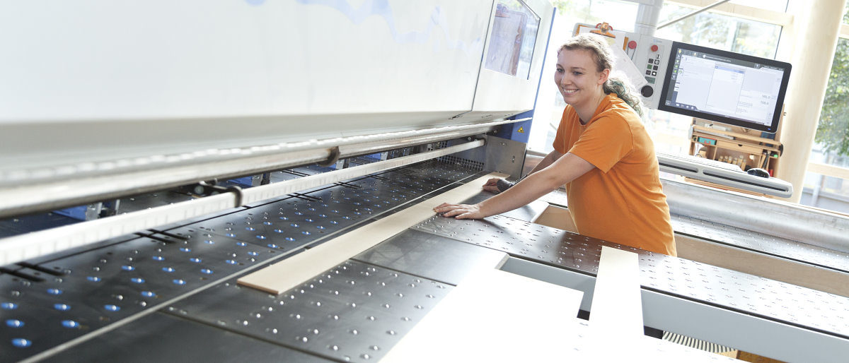 Aktuelle Zahlen und Tipps zum Ausbildungsstart in der Region. Eine junge Frau legt in einer Werkstatt lächelnd eine Holzlatte in eine moderne CNC-Maschine.