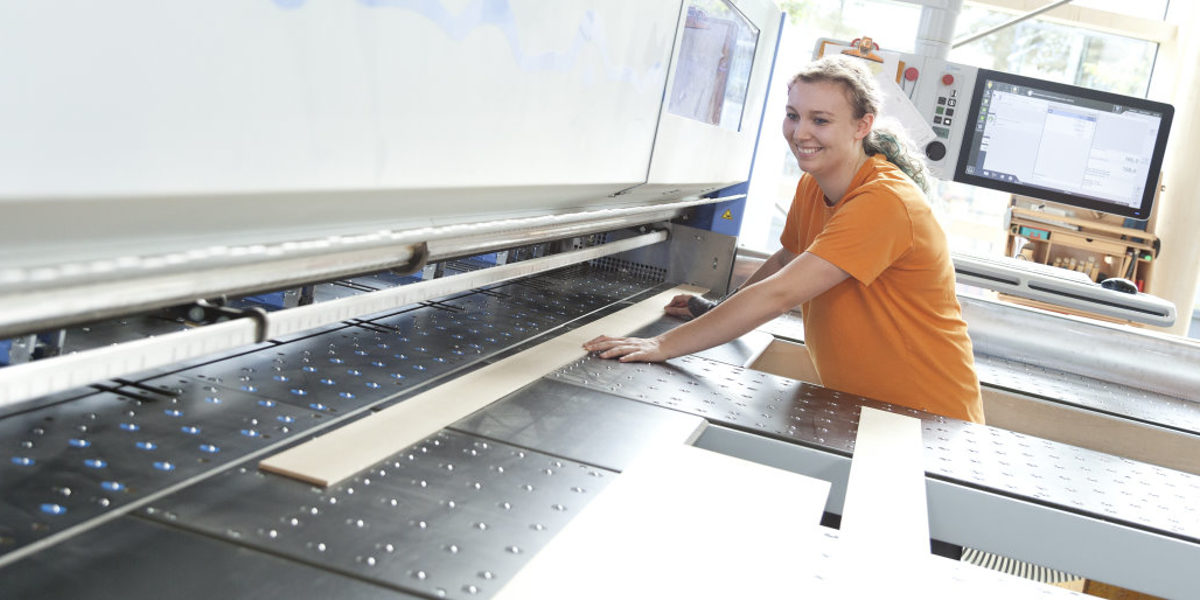 Eine junge Frau legt in einer Werkstatt lächelnd eine Holzlatte in eine moderne CNC-Maschine.