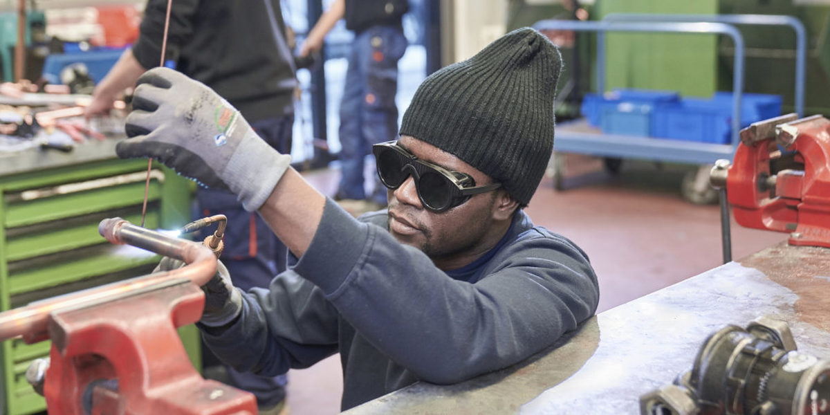 Ein Mann mit dunkler Hautfarbe, Mütze und Schutzbrille bearbeitet in einer Werkstatt ein Metallrohr mit einem Schweißgerät.