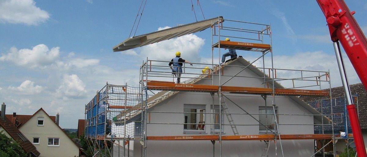 Ein Teil des Daches eines Fertighauses wird mit dem Kran auf den Rohbau gesetzt. Zwei Bauarbeiter stehen auf dem Gerüst.