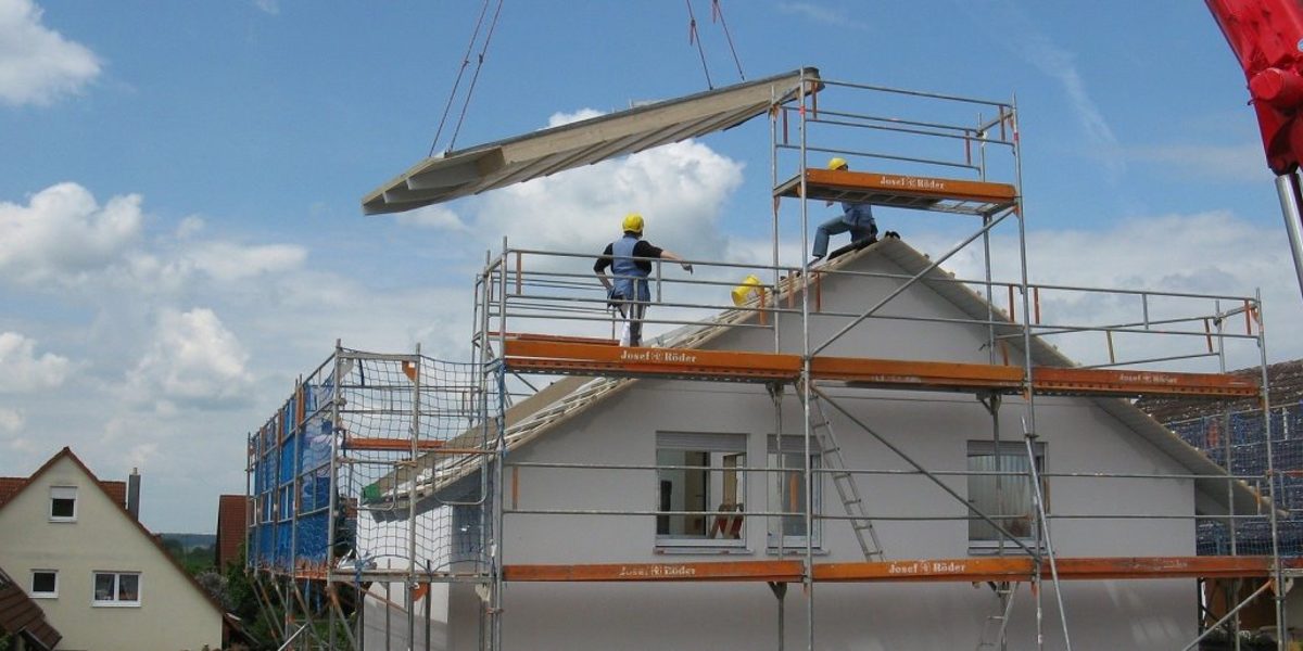Ein Teil des Daches eines Fertighauses wird mit dem Kran auf den Rohbau gesetzt. Zwei Bauarbeiter stehen auf dem Gerüst.