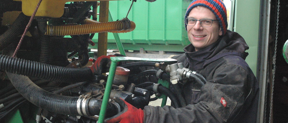 Land- und Baumaschinenmechatroniker Franz Kornder ist PLW-Landessieger Ein Mann mit Brille in Mütze, Winterjacke und Handschuhen greift arbeitet an einer Landmaschine und lächelt in die Kamera.