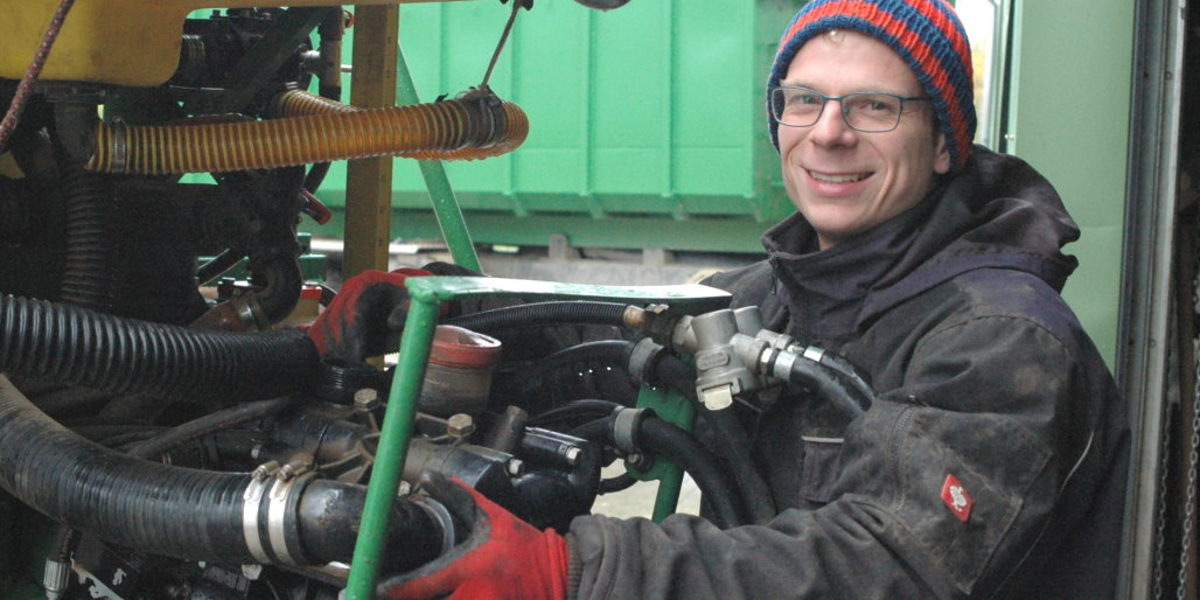 Ein Mann mit Brille in Mütze, Winterjacke und Handschuhen greift arbeitet an einer Landmaschine und lächelt in die Kamera.