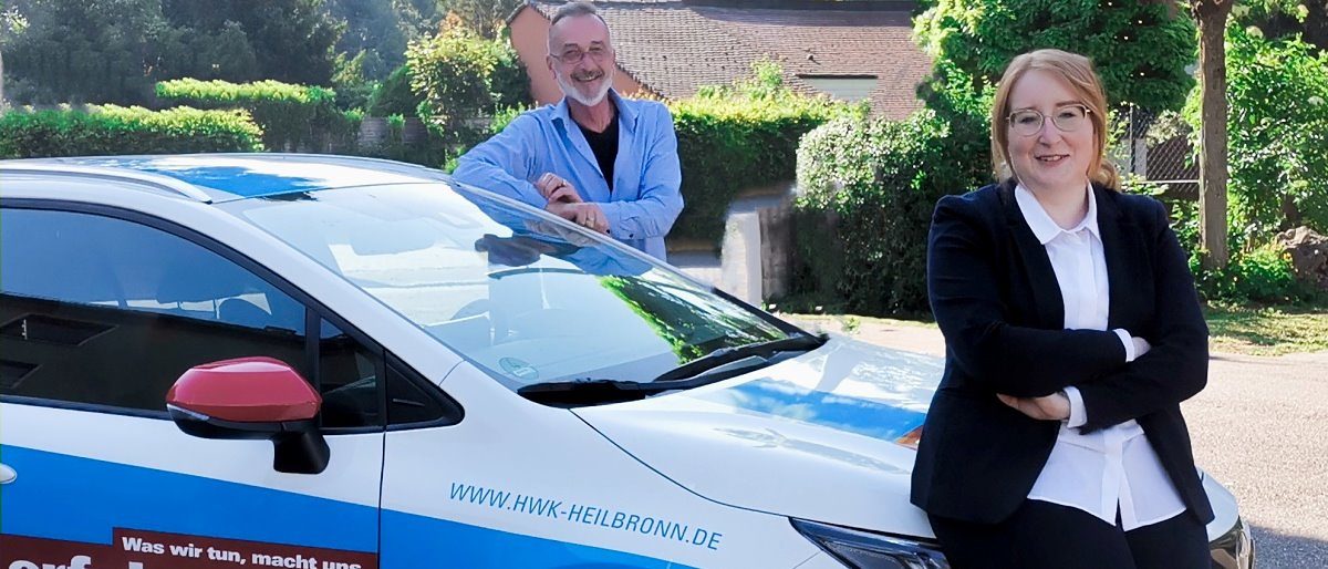 Peter Bauer und Lisa Büchele sind Berater der Passgenauen Besetzung. Ein Auto auf einem Parkplatz. An der Motorhaube steht angelehnt eine Frau, an der Fahrertür ein Mann, lächeln in die Kamera.