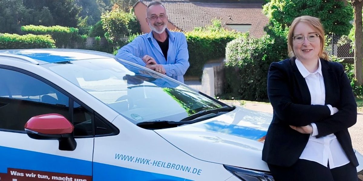 Peter Bauer und Lisa Büchele sind Berater der Passgenauen Besetzung. Ein Auto auf einem Parkplatz. An der Motorhaube steht angelehnt eine Frau, an der Fahrertür ein Mann, lächeln in die Kamera.
