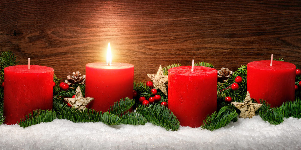 Adventsgesteck mit Tannenzweigen und vier roten Kerzen, eine Kerze brennt.
