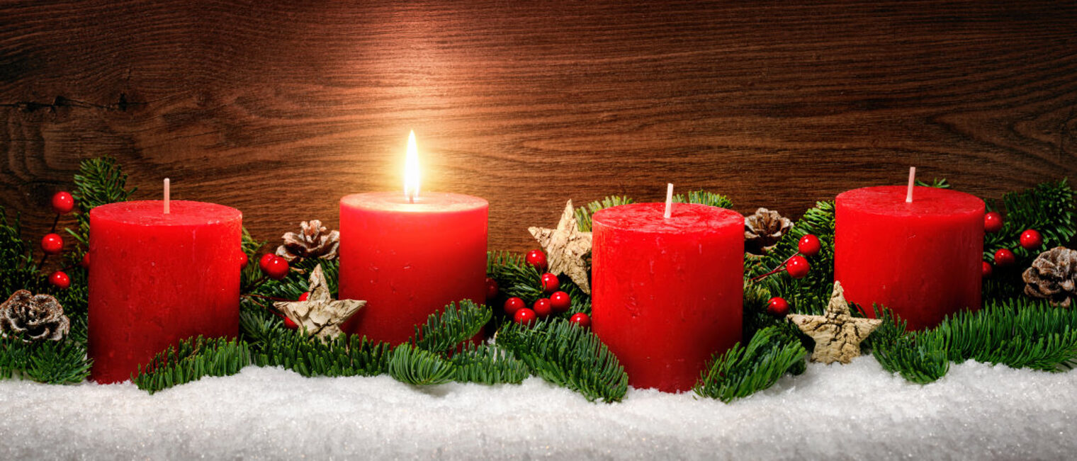 Adventsgesteck mit Tannenzweigen und vier roten Kerzen, eine Kerze brennt.