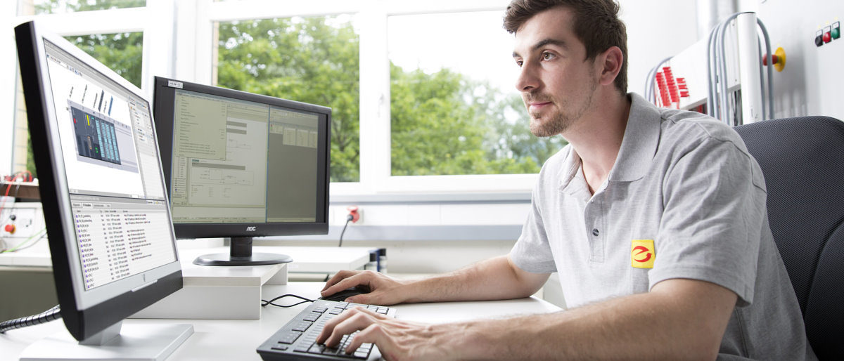 Ein junger Mann sitzt in einem Büro an einem Schreibtisch und blickt konzentriert auf einen von zwei Monitoren.