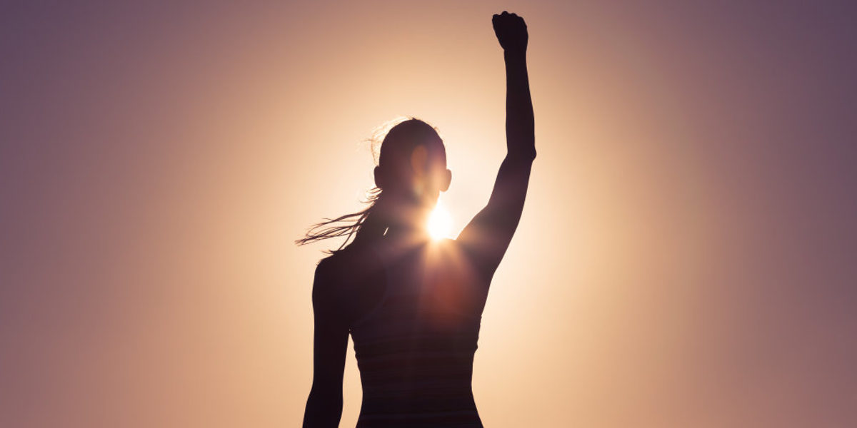 Die Silhoutte einer langhaarigen Frau, die vor der Sonne steht und den rechten Arm in einer Siegerpose nach oben gereckt hat.