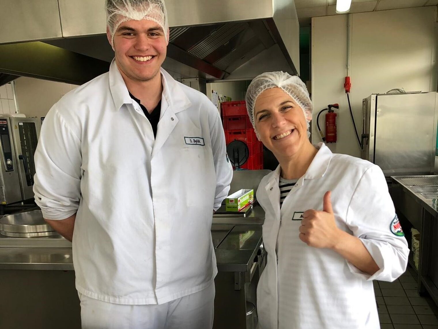 Ein junger Mann und eine Frau stehen in weißer Arbeitskleidung und mit Haarnetz in einer Profi-Küche und lachen in die Kamera