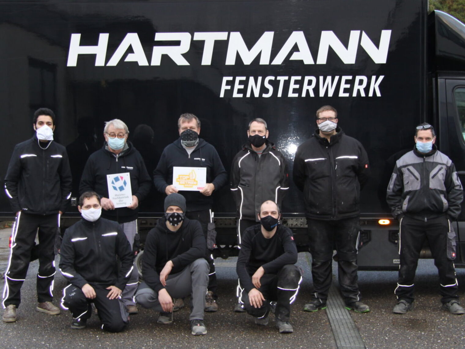 Eine Gruppe von neuen Männern in Arbeitskleidung steht bzw. kniet vor einem schwarzen Transporter mit weißem Schriftzug.