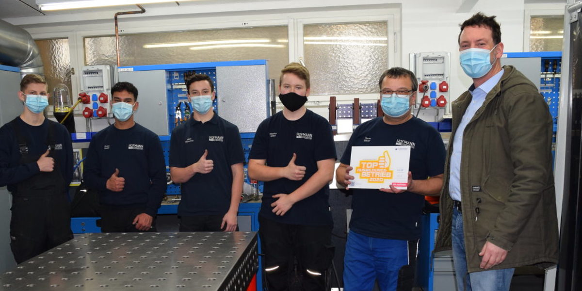 Sechs Männer stehen in einer Werkstatt. Alle tragen Mund-Nasen-Masken. Einer hält ein Schild mit einem Logo vor sich.