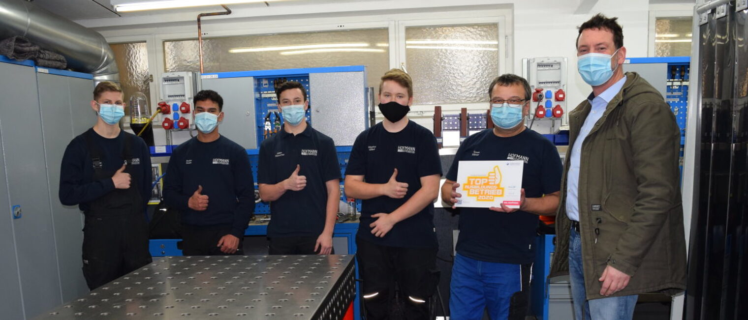 Die Hofmann GmbH in Blaufelden wurde zum 2. Mal Top-Ausbildungsbetrieb Sechs Männer stehen in einer Werkstatt. Alle tragen Mund-Nasen-Masken. Einer hält ein Schild mit einem Logo vor sich.