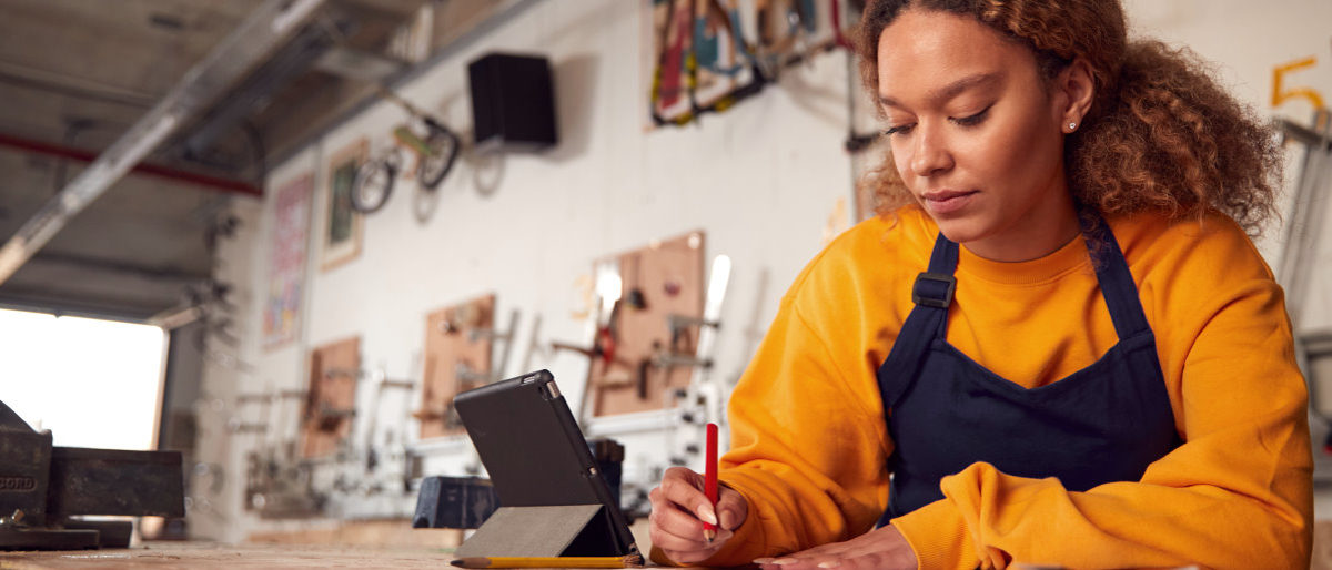 Online-Speed-Dating für Handwerksbetriebe und Ausbildungssuchende. Eine junge Frau sitzt an einem Tisch in einer Werkstatt. Neben ihr steht ein Tablet. Sie hat einen Stift in der Hand.