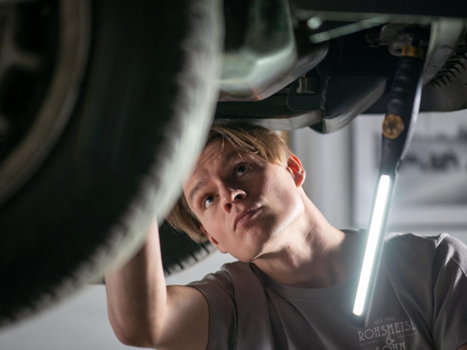 Ein junger Mann blickt prüfend unter eine Auto auf einer Hebebühne mit einer Arbeitsleuchte in der Hand.