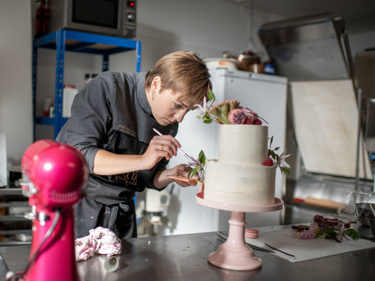 Eine Frau mit kurzen Haaren arbeitet an einer zweistöckigen Torte in einer professionellen Küche.