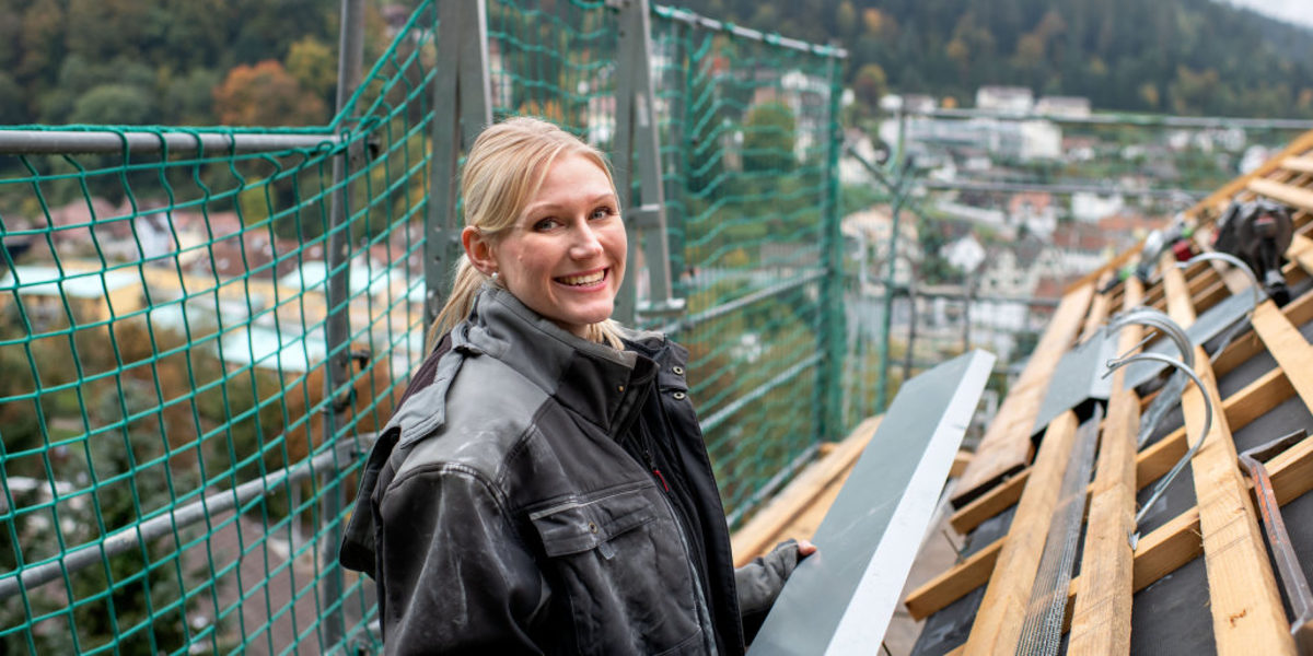 Spenglerin Luisa Buck ist eine von vier Botschaftern für das Handwerk. Eine blonde junge Frau steht in Arbeitskleidung auf einem Gerüst vor einem ungedeckten Dach in der Hand ein langes Blechstück