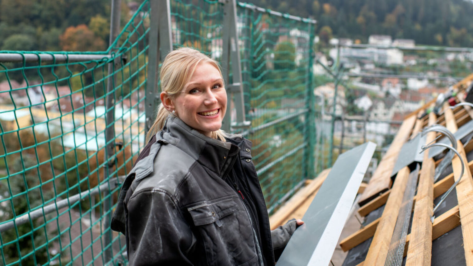 Eine blonde junge Frau steht in Arbeitskleidung auf einem Gerüst vor einem ungedeckten Dach in der Hand ein langes Blechstück