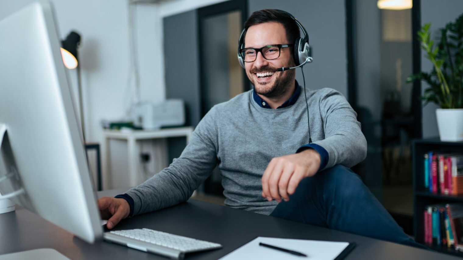 Mann mit Brille und Headset sitzt in einem Büro an einem Schreibtisch vor dem Computer und lacht in Richtung des Monitors.