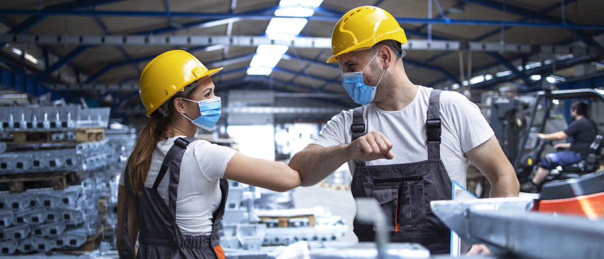 Betriebe können Corona-Vorschriften wieder weitestgehend selbst regeln Eine Frau und ein Mann in Arbeitskleidung mit Bauhelm und Maske begrüßen sich mit dem Ellenbogen in einer Fabrikhalle.