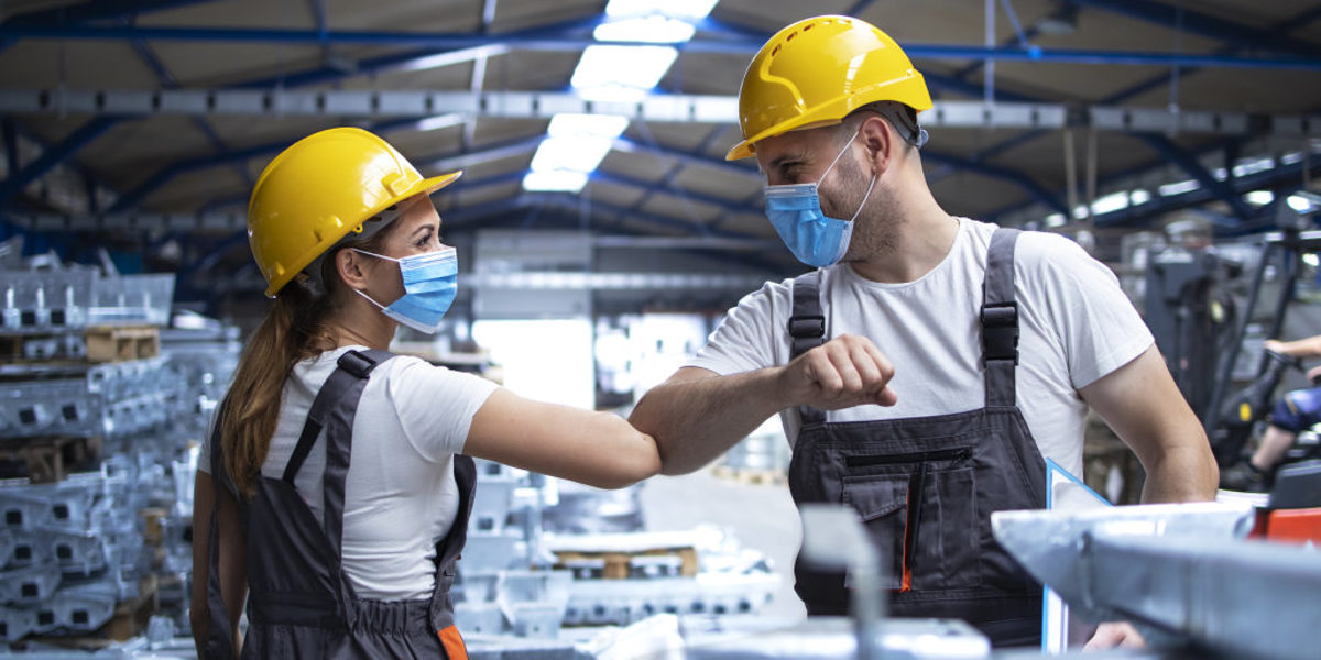 Eine Frau und ein Mann in Arbeitskleidung mit Bauhelm und Maske begrüßen sich mit dem Ellenbogen in einer Fabrikhalle.