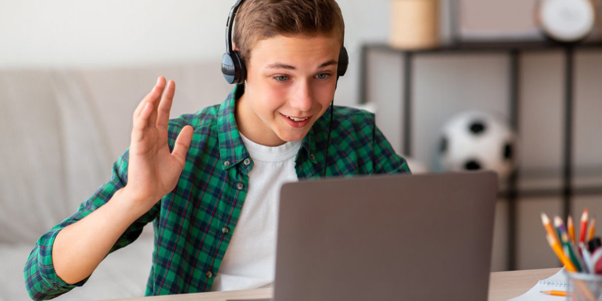 Ein Junge in kariertem Hemd und T-Shirt sitzt am Tisch vor offenem Laptop mit Kopfhörern und winkt in Richtung Bildschirm.