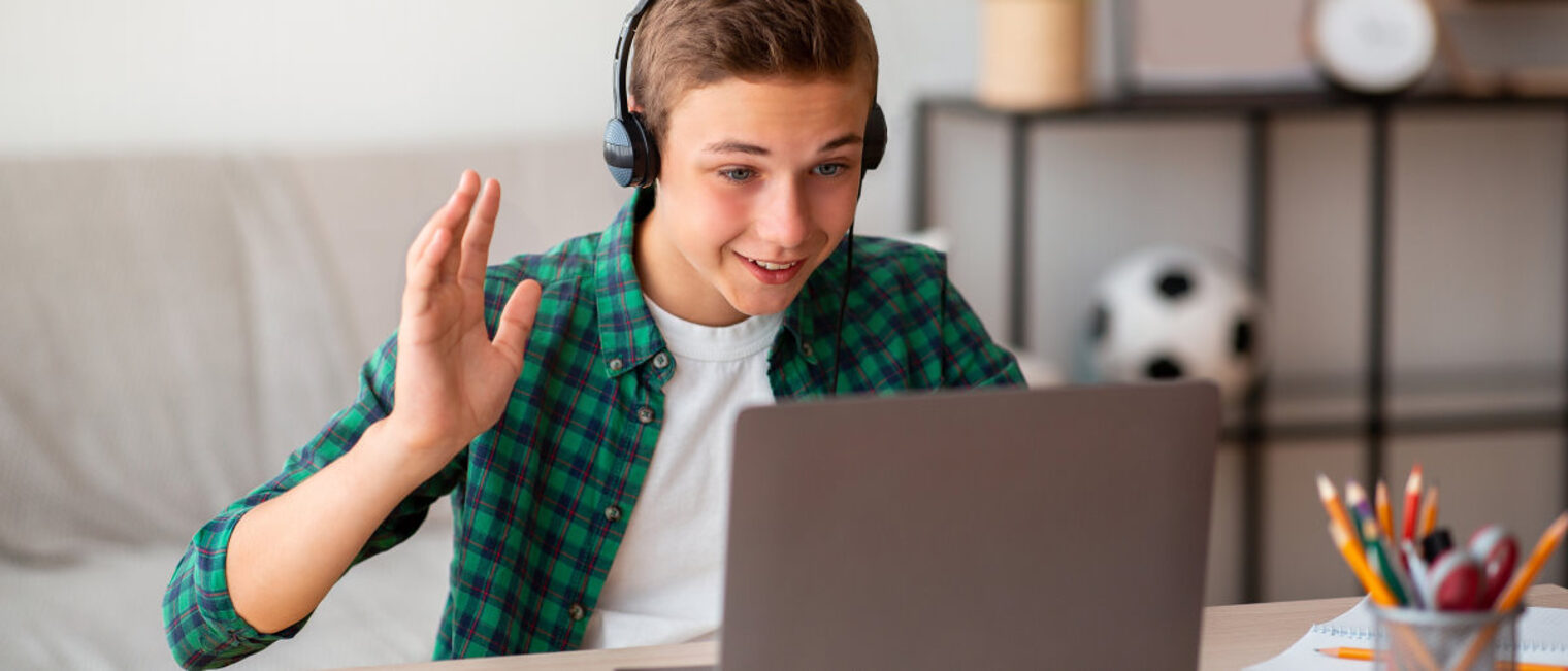 Ein Junge in kariertem Hemd und T-Shirt sitzt am Tisch vor offenem Laptop mit Kopfhörern und winkt in Richtung Bildschirm.