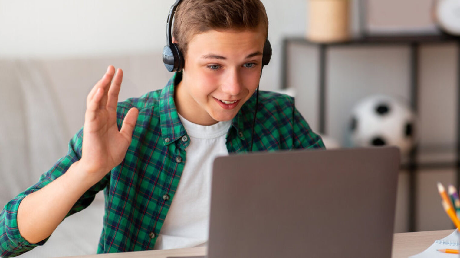 Ein Junge in kariertem Hemd und T-Shirt sitzt am Tisch vor offenem Laptop mit Kopfhörern und winkt in Richtung Bildschirm.