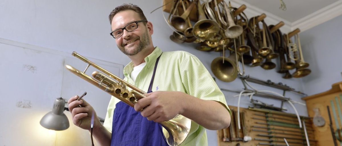 Handwerker beweisen auch in der Pandemie, wie kreativ sie sind. Ein Mann hält in der einen Hand eine Trompete, in der anderen ein Werkzeug. Hinter ihm hängen weitere Instrumente an der Wand.