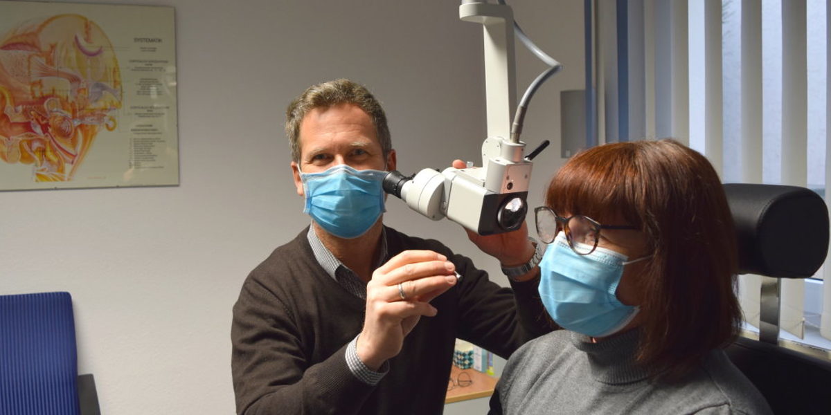 Hörakustiker versorgen auch im Lockdown ihre Kunden. Ein Mann mit Mundschutz sitzt an einem Gerät zur Untersuchung des Gehörs einer Frau mit Mundschutz und Brille.